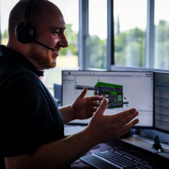 Technischer Experte, der über ein Headset Fernsupport für Kunden leistet und dabei mit einer digitalen Maschinenschnittstelle interagiert, die auf drei Monitoren angezeigt wird.